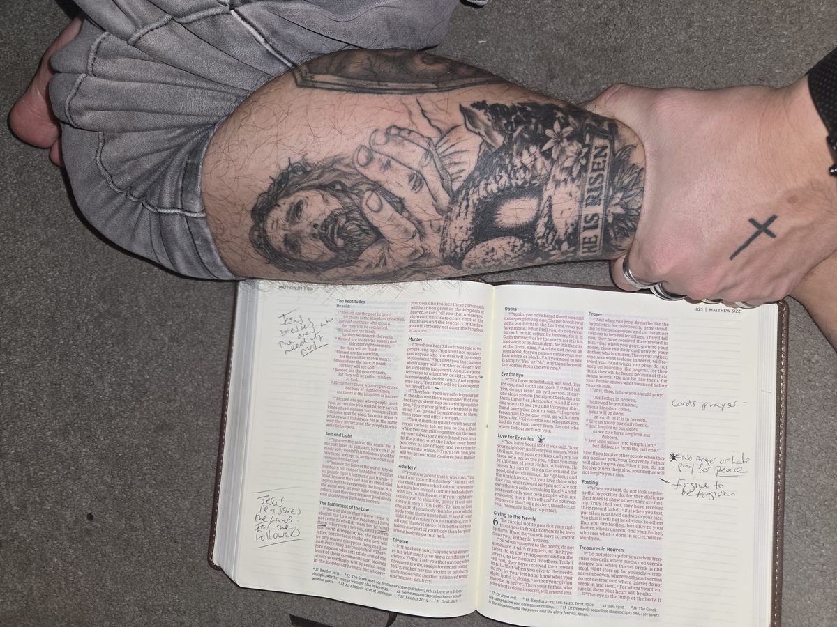 A tattoo of Jesus with the words He is risen, beside an open Bible at Matthew 5 and 6 with handwritten notes in the margins